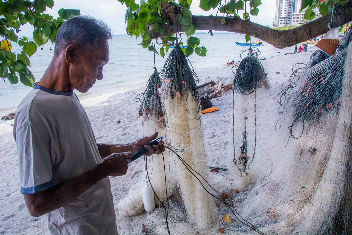 Mr Kamaruddin, 70, fixes his net on the beach next to his home at Tanjung Tokong, Penang Island. Credit: Rod Harbinson via Mongabay